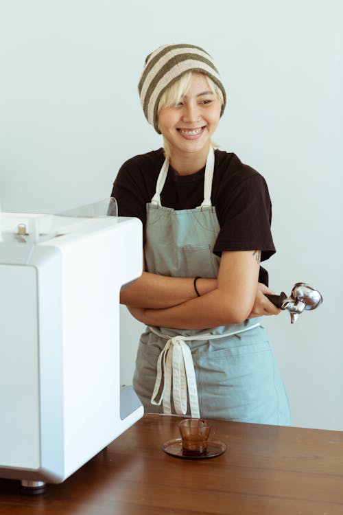 Positive young Asian female worker in apron with portafilter in hand laughing while standing near table with coffee machine