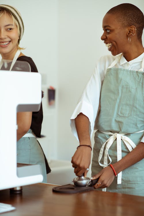 Optimistic laughing diverse female baristas wearing aprons pressing ground coffee in portafilter while working in modern light kitchen and chatting