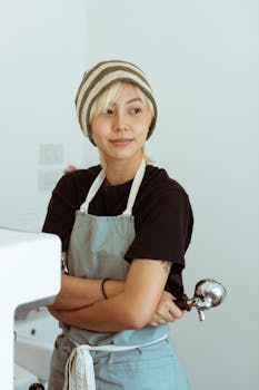 Young female barista with arms crossed in a contemporary café, holding a portafilter and looking confident.