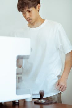 Crop focused Asian male barista in casual white shirt using professional coffee machine and barista tools in light kitchen