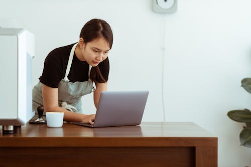 Concentrated young Asian lady in casual black shirt and apron with cup of hot drink watching cooking tutorials via modern netbook on wooden table against white wall
