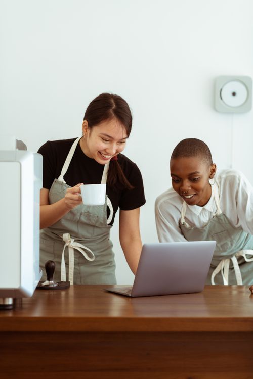 Free Positive joyful multiethnic waitresses in casual wear and aprons browsing laptop while gathering together at table with cup of beverage in light modern kitchen Stock Photo