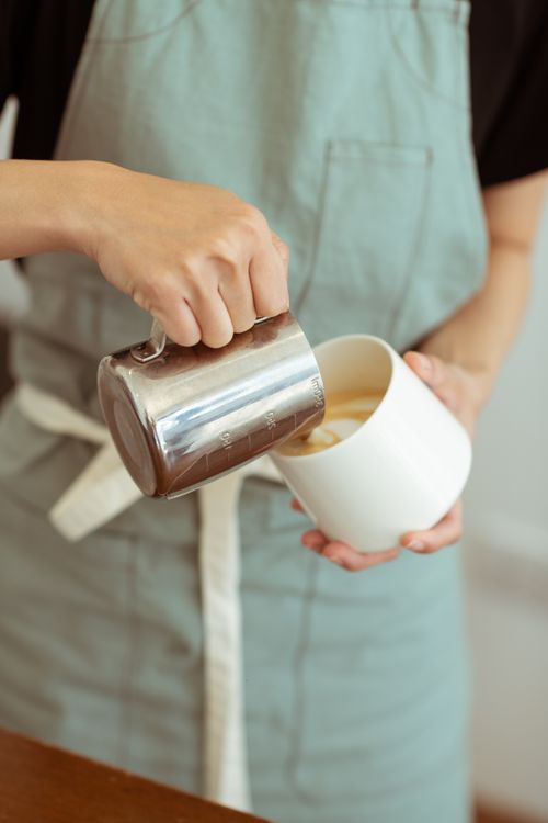 Free Crop anonymous female barista in light green apron preparing fresh cappuccino and pouring steamed milk into white ceramic cup Stock Photo