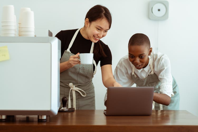 Happy Colleagues In Aprons Browsing Laptop In Kitchen