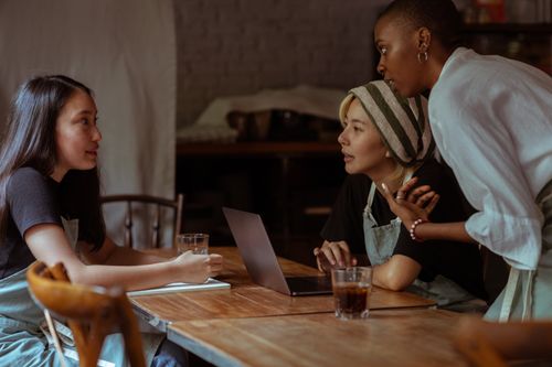 Free Serious multiethnic waitresses in aprons sitting at wooden table with modern netbook and notebook while discussing business project Stock Photo