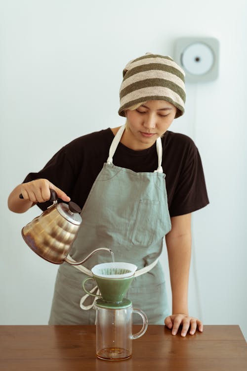 Focused Asian female barista wearing hat and apron preparing delicious coffee alternatively by pouring hot water from kettle into pour over while working in light kitchen