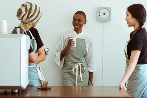 Positive smiling diverse female cafe workers in aprons gathering together in light kitchen with cup of hot drink and chatting during coffee break