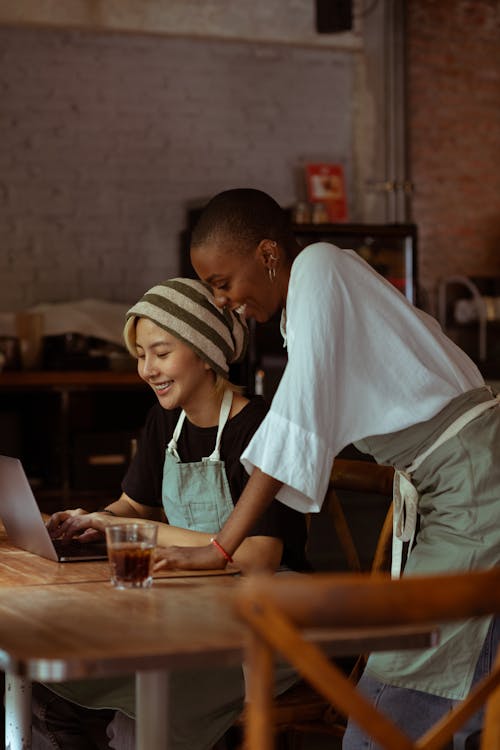 Side view of positive diverse female colleagues in aprons surfing modern netbook while gathering at wooden table with drink in modern cafe