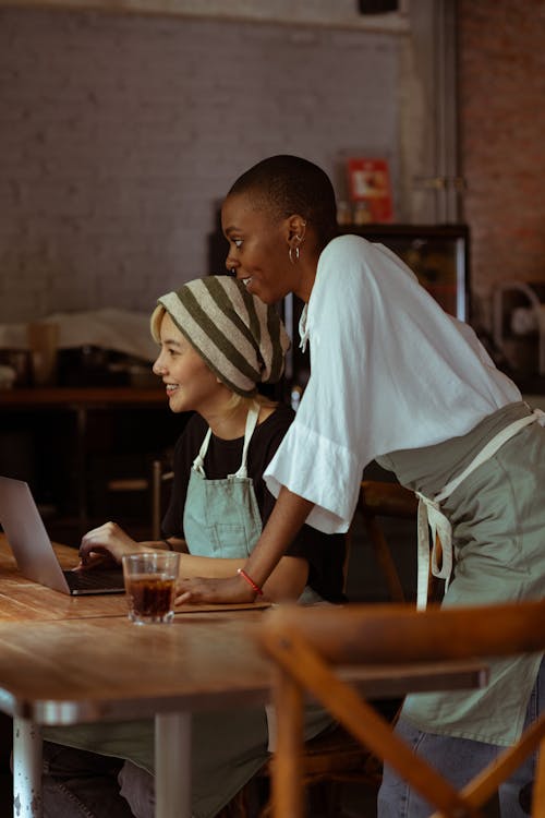 Side view of positive smiling diverse female cafe workers in casual wear and aprons working on contemporary netbook while gathering at table and looking away happily