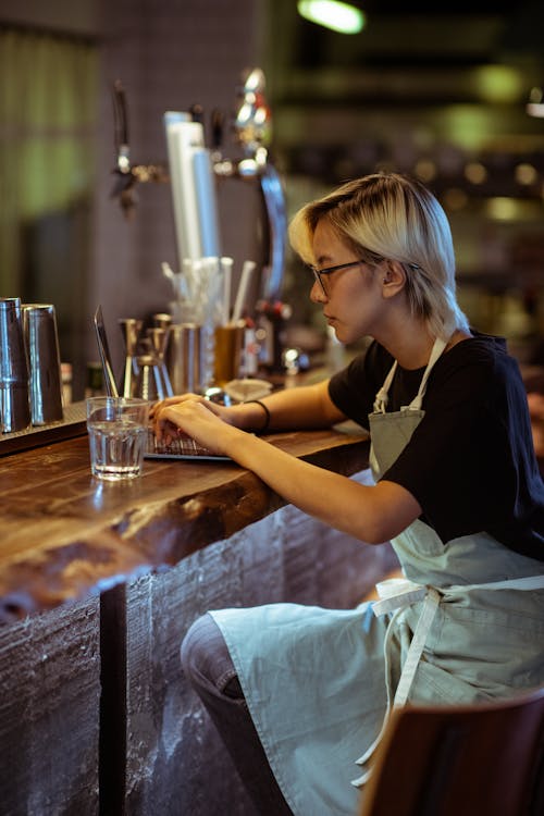 Free Side view of serious bartender in apron and eyeglasses using modern netbook while sitting at bar counter in modern restaurant Stock Photo