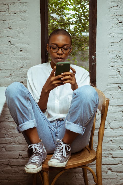 Free Full length of young black concentrated female in jeans and white blouse sitting on wooden chair against narrow window and white brick wall while surfing smartphone Stock Photo