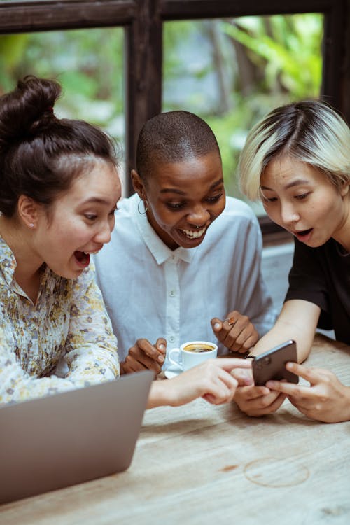 Free Amazed surprized multiethnic female friends in casual clothes with cup of coffee browsing smartphone while sitting at wooden table with laptop against window Stock Photo