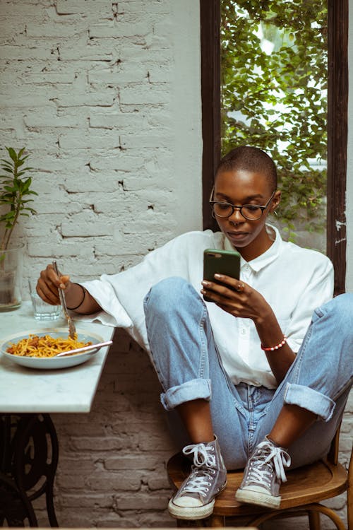 Calm thoughtful young African American lad in casual clothing sitting at table while surfing modern smartphone and eating yummy pasta against white brick wall and window