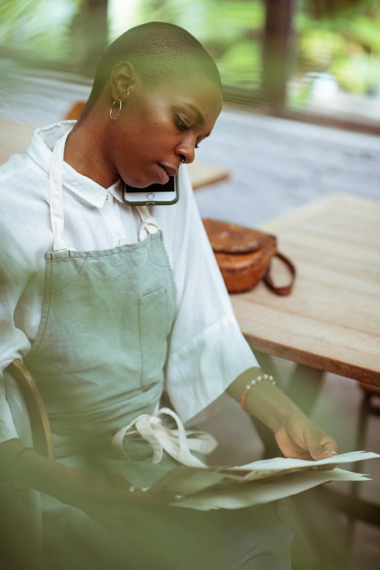 Attentive Black Waitress Talking On Smartphone And Looking At Papers