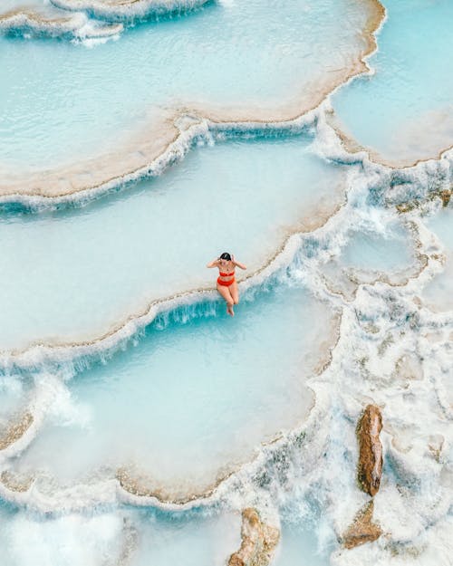 Ücretsiz Kırmızı Bikinili Kadın Sırasında Su Gövdesinin önünde Kahverengi Kaya Oluşumu üzerinde Duran Stok Fotoğraflar