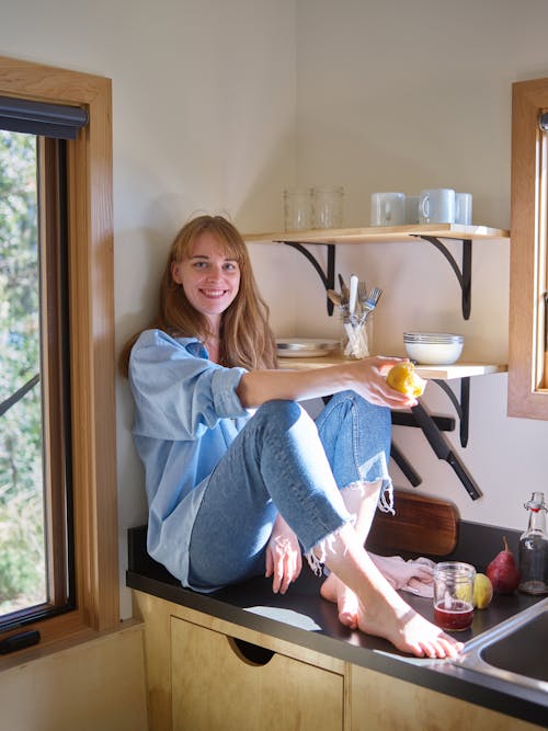 Free Full length young slim female in denim sitting on modern kitchen cabinet with apple on sunny day and looking at camera Stock Photo