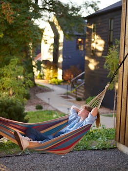 Full body of content blonde wearing casual clothes relaxing in striped hammock in rural village on sunny summer day with the Quote "Never complain and never explain." written on it and have average color value #68614D