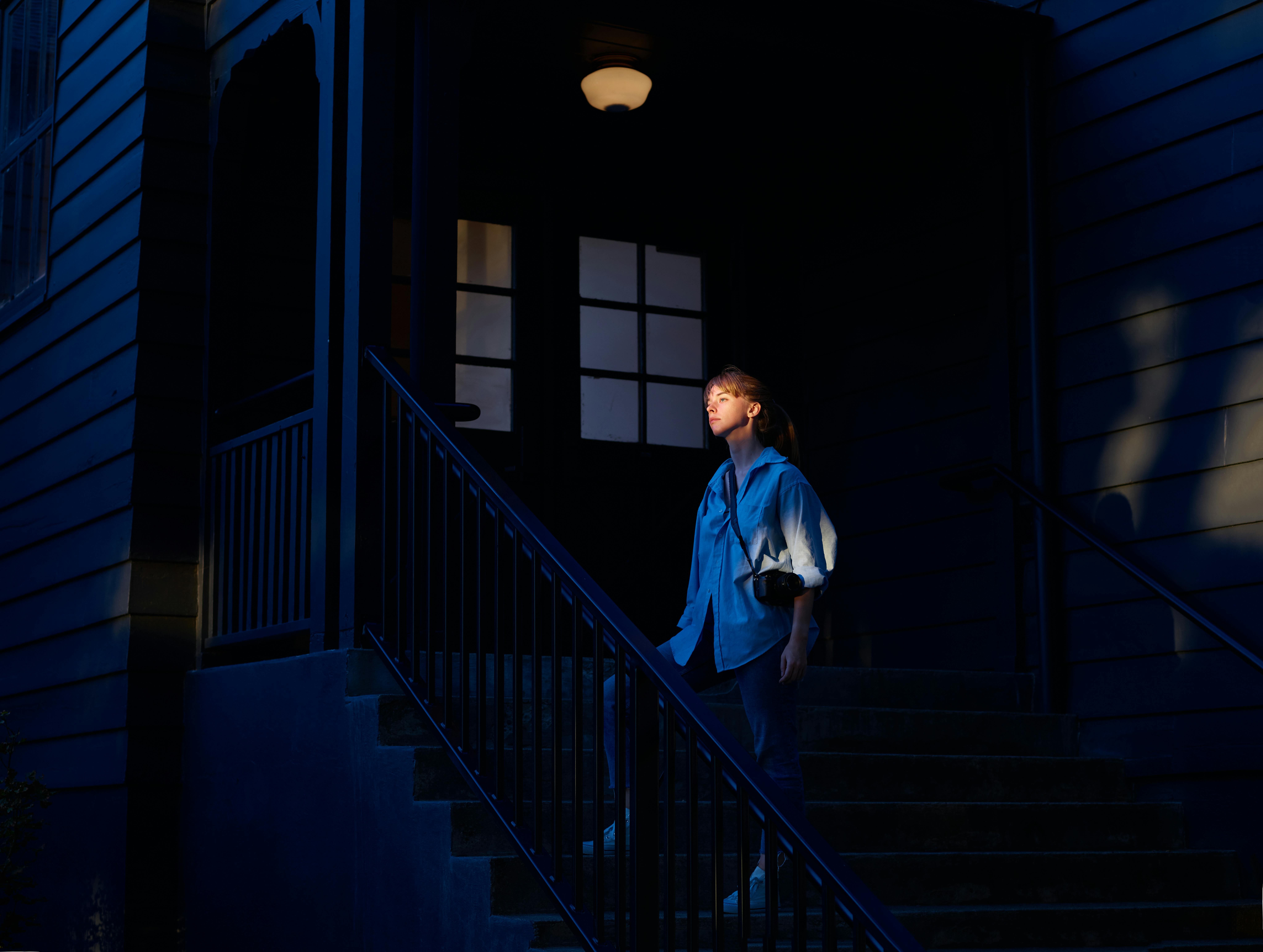 Full body calm pensive female in blue casual wear standing on staircase leading to cottage entrance at dark night and looking away