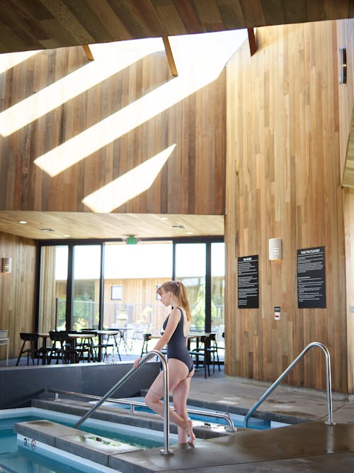 Free Side view full body unrecognizable slim gorgeous female wearing black swimsuit standing near indoors swimming pool railing on sunny day Stock Photo