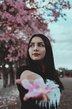 春天，公园里一位身穿黑色服装的宁静女子，手持粉红色的花朵，身旁是盛开的树木