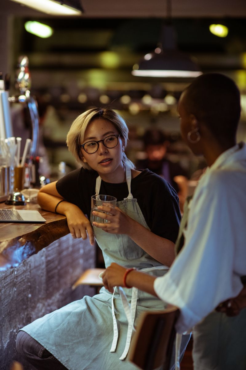 Positive diverse female bartenders wearing aprons sitting at bar counter