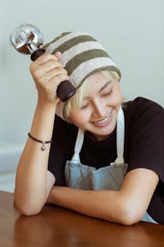 Happy smiling Asian female barista wearing apron and striped hat sitting at table at leaning on hand with portafilter