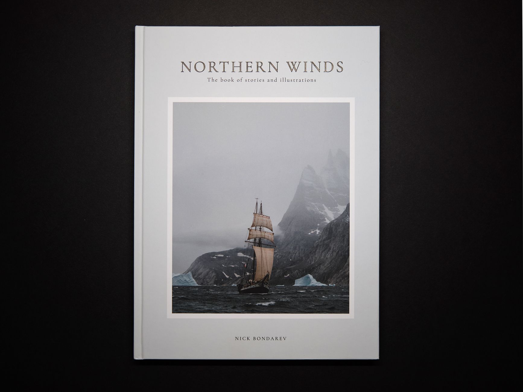 Modern interesting paper book telling about north seas traveling in hardcover with picture of old fashioned ship with white sails near rocky snowy cliff in thick fog on black background