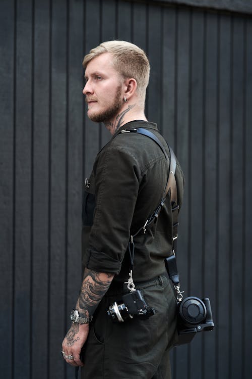 Side view of serious young bearded male photographer in black stylish outfit wearing belt with professional photographer equipment and looking away thoughtfully while standing against black plank wall