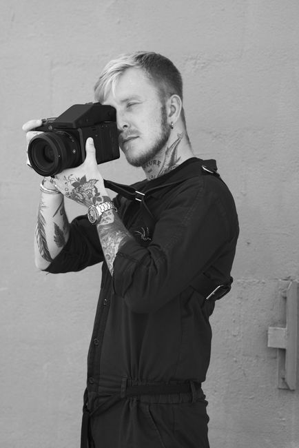 Black and white side view confident bearded photographer in stylish black clothes taking shots on contemporary professional photo camera while standing against gray background