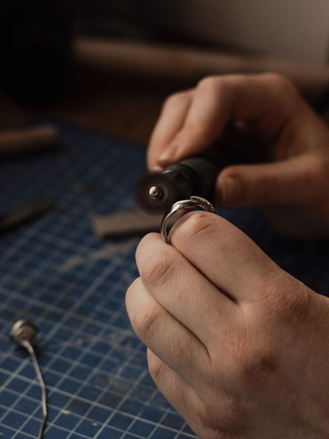 Two hands finishing a plain silver ring with a rotary tool on a blue cutting mat at a workbench