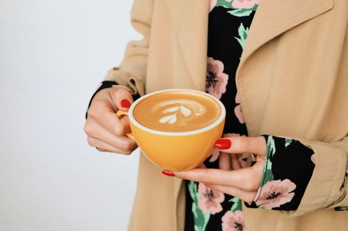 Free Close-up of a woman holding a cappuccino with latte art, showing red nails and floral dress. Stock Photo