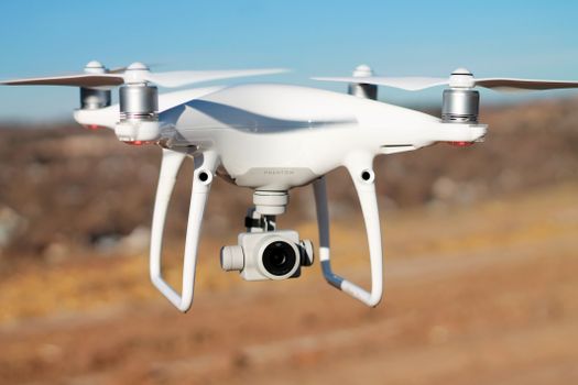 A white drone with camera hovering outdoors during the day against a blurred landscape backdrop.