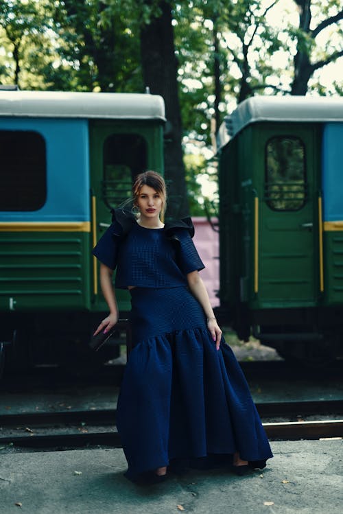 Full length of young elegant female traveler in maxi dress sitting on suitcase and looking at camera near retro train on railway platform