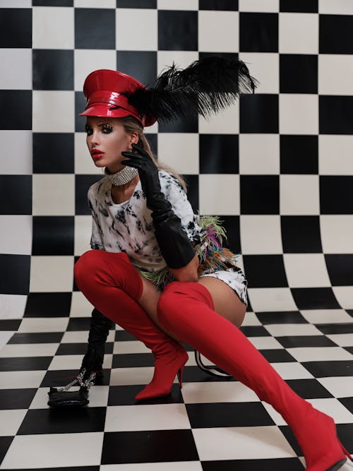 Side view of self confident young female in red boots and hat sitting on haunches in checkered room and looking at camera