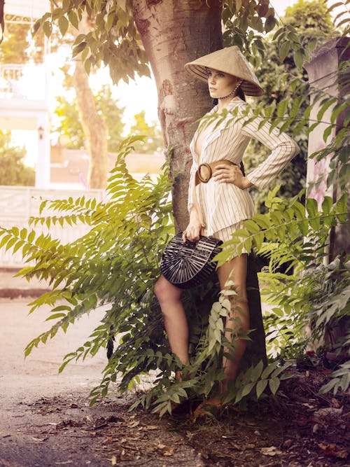 Side view of stylish young woman in classy outfit and Vietnamese straw hat standing near green tree with hand on waist and looking away