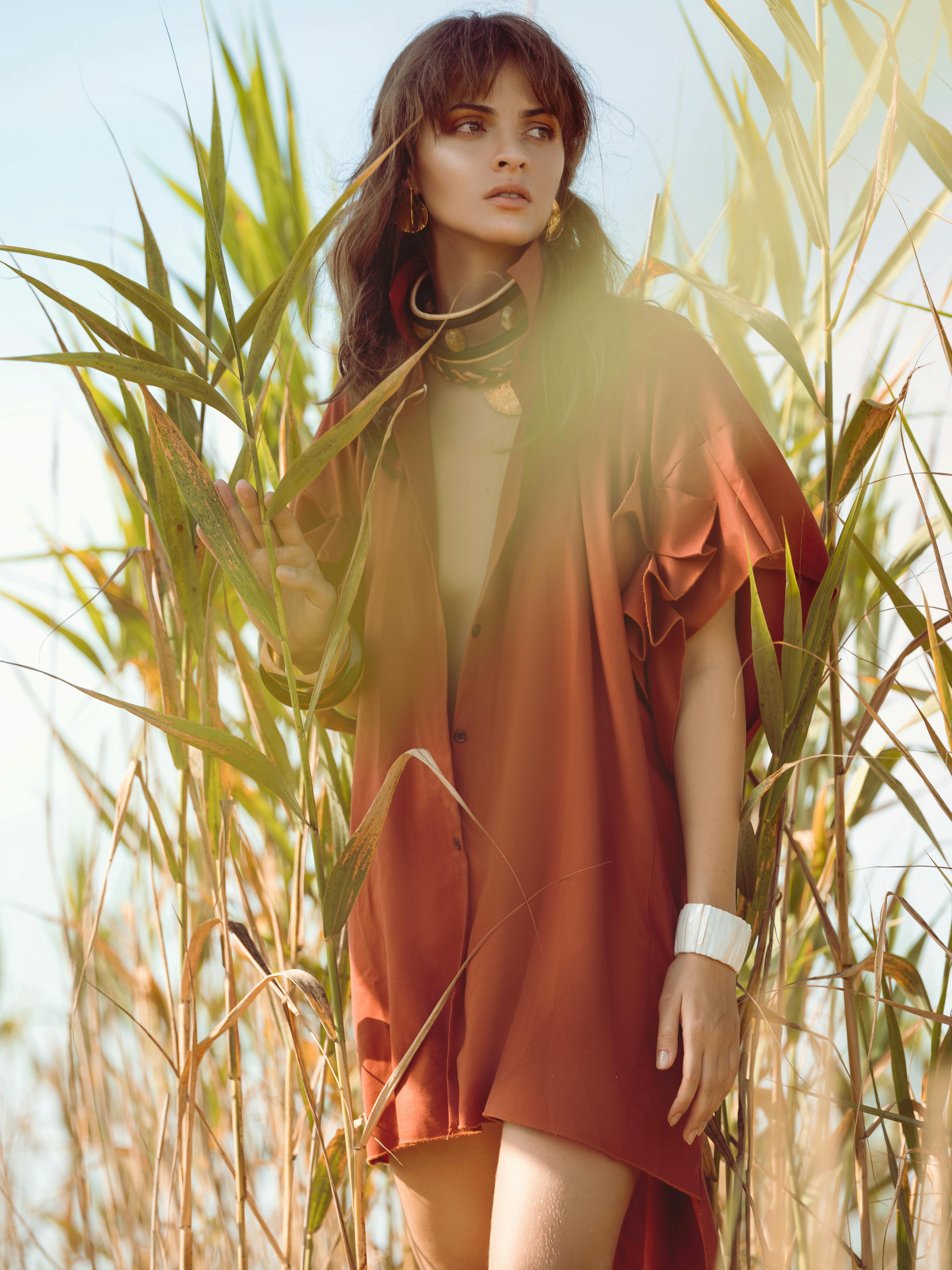 Calm young female traveler in stylish sundress and ethnic accessories standing near tall green grass in field and looking away