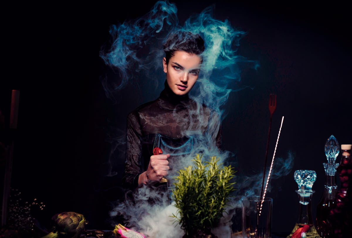 Graceful young female alchemist with knife in hand in black outfit preparing potion from various herbs among smoke in dark room