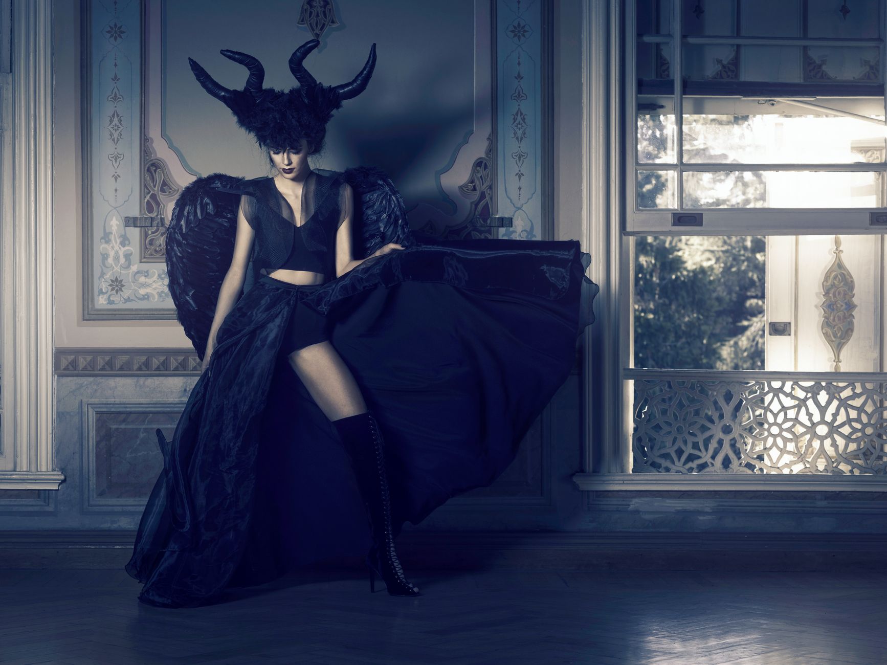 A dramatic editorial fashion portrait of a model in black with horns, captured in a moody interior setting.