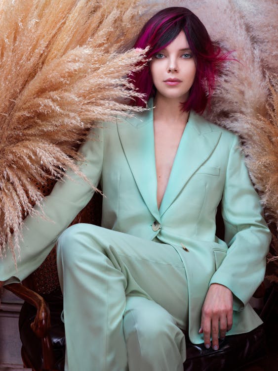 Fashionable young female with bright hair sitting on chair with legs crossed among dried plants