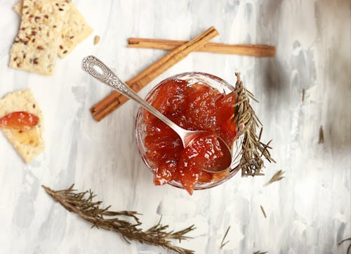 Top view composition of bowl with delicious jam in cup with rosemary sprigs and crispbread with cinnamon sticks