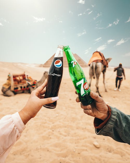 Unrecognizable crop tourists clinking glass bottles of soda while travelling in sandy desert