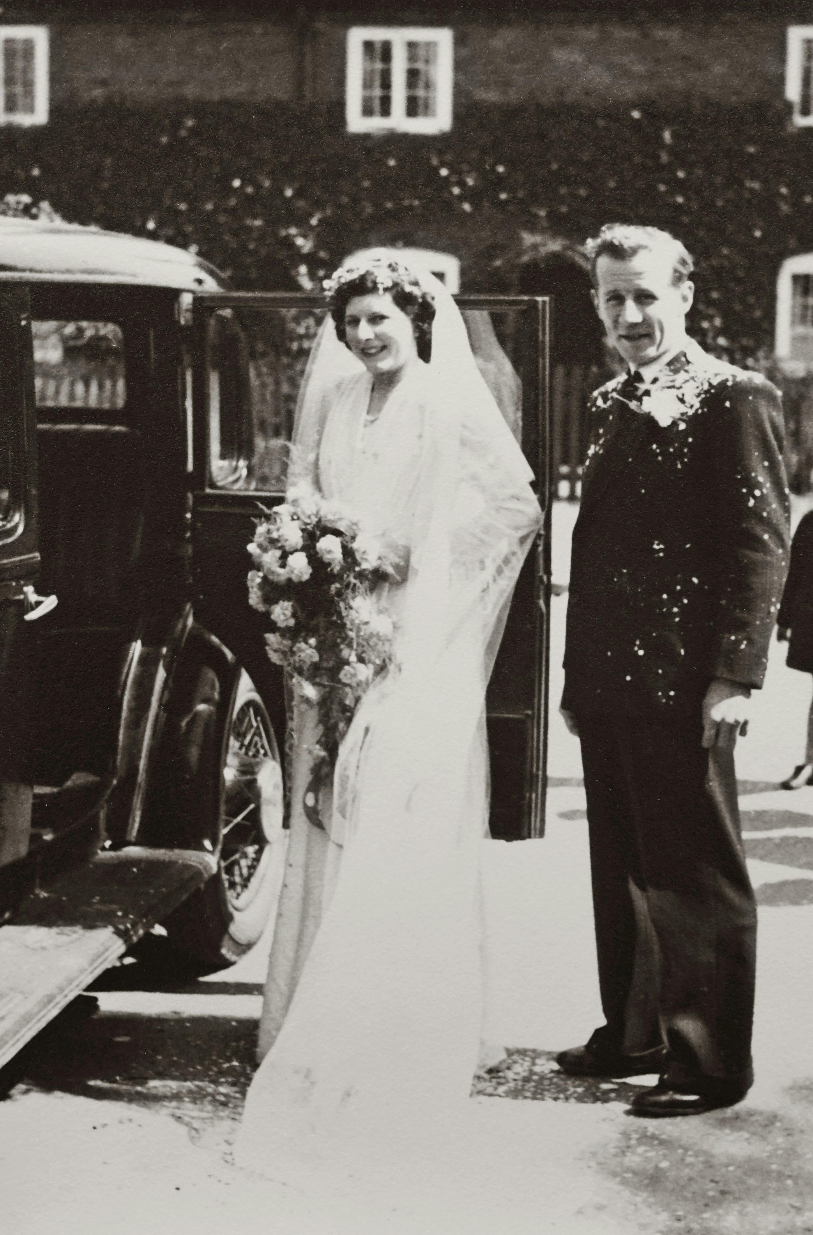 A timeless black and white portrait of a bride and groom standing by a classic car on their wedding day.