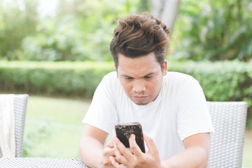 Concentrated ethnic male with stylish haircut wearing casual white shirt sitting on chair on sunny veranda and browsing cellphone