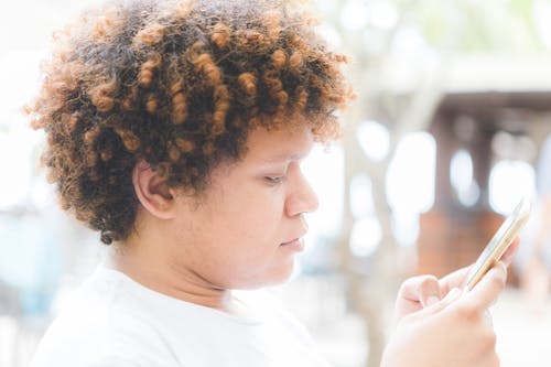 Free Side view crop concentrated ethnic male with curly hair browsing contemporary mobile phone while spending sunny day on veranda Stock Photo