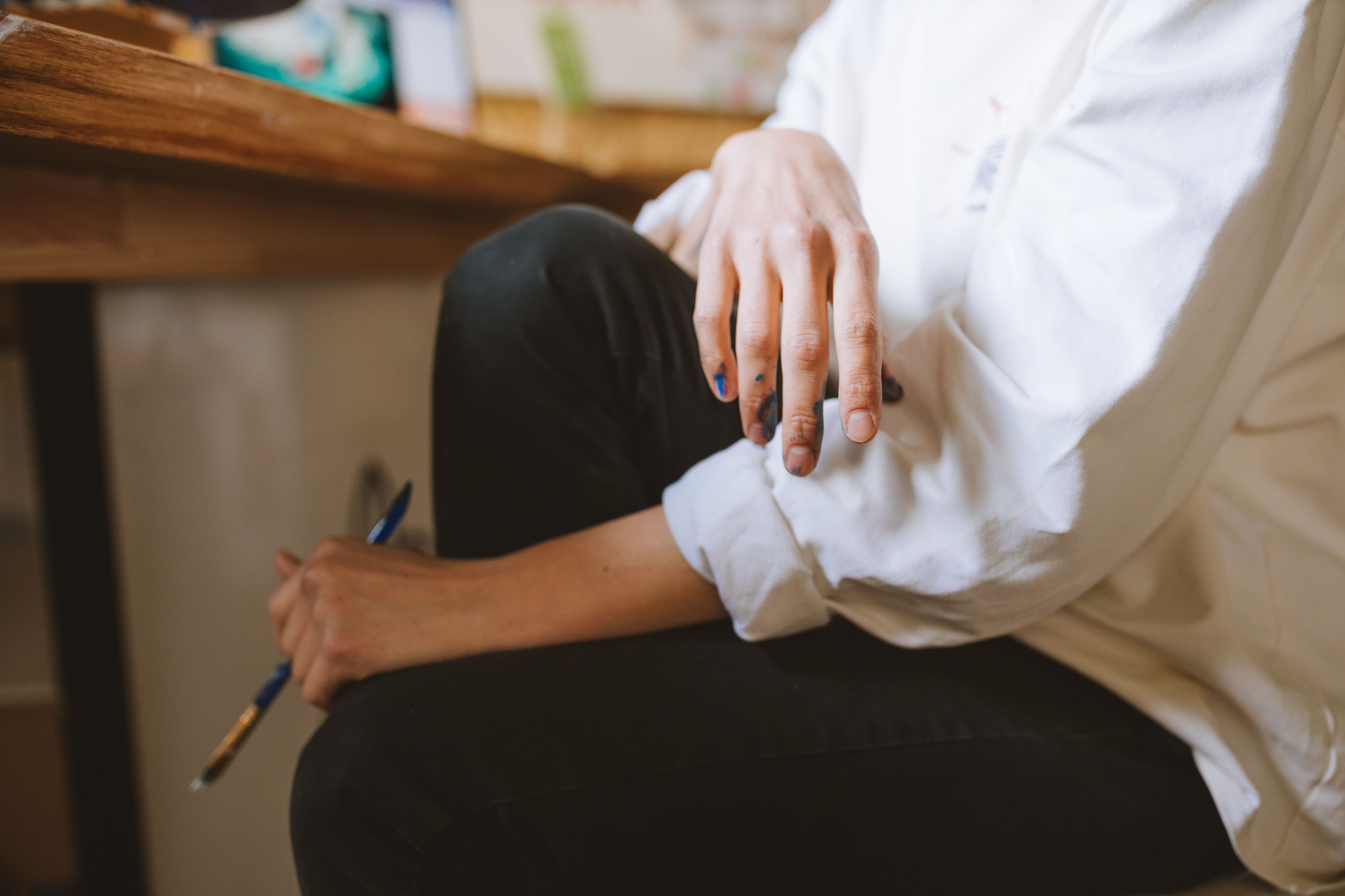 A close-up view of a painter's hand with paint marks holding a brush beside a wooden table.