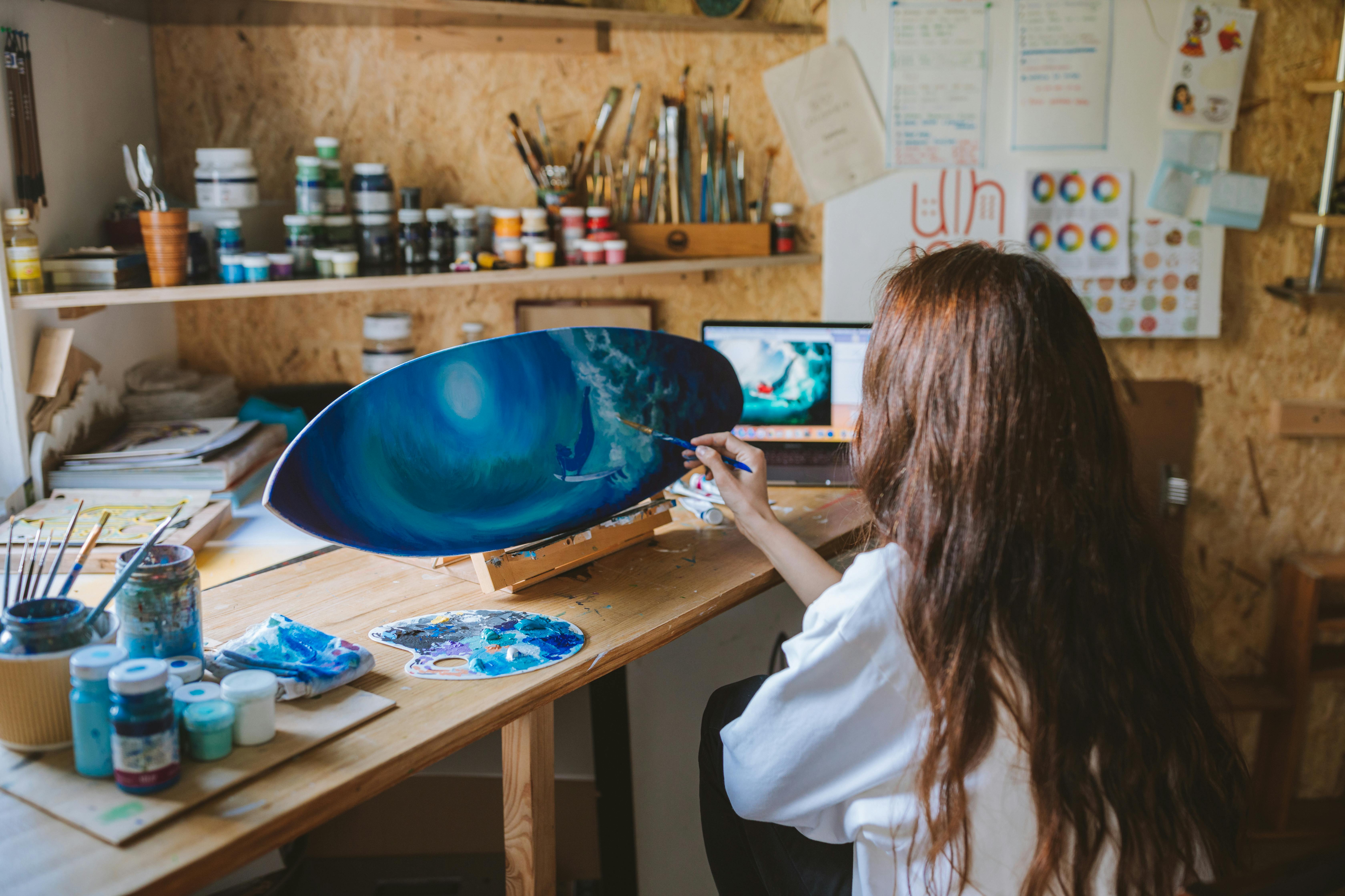 A woman artist painting a skateboard in a well-organized home studio with vibrant materials.