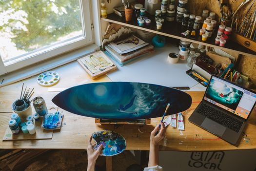 An artist paints a surfboard in a vibrant workshop setting, surrounded by paint supplies and a laptop.