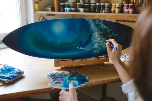 Free Artist painting a surf scene on a surfboard, showcasing artistic creativity in a sunlit studio. Stock Photo