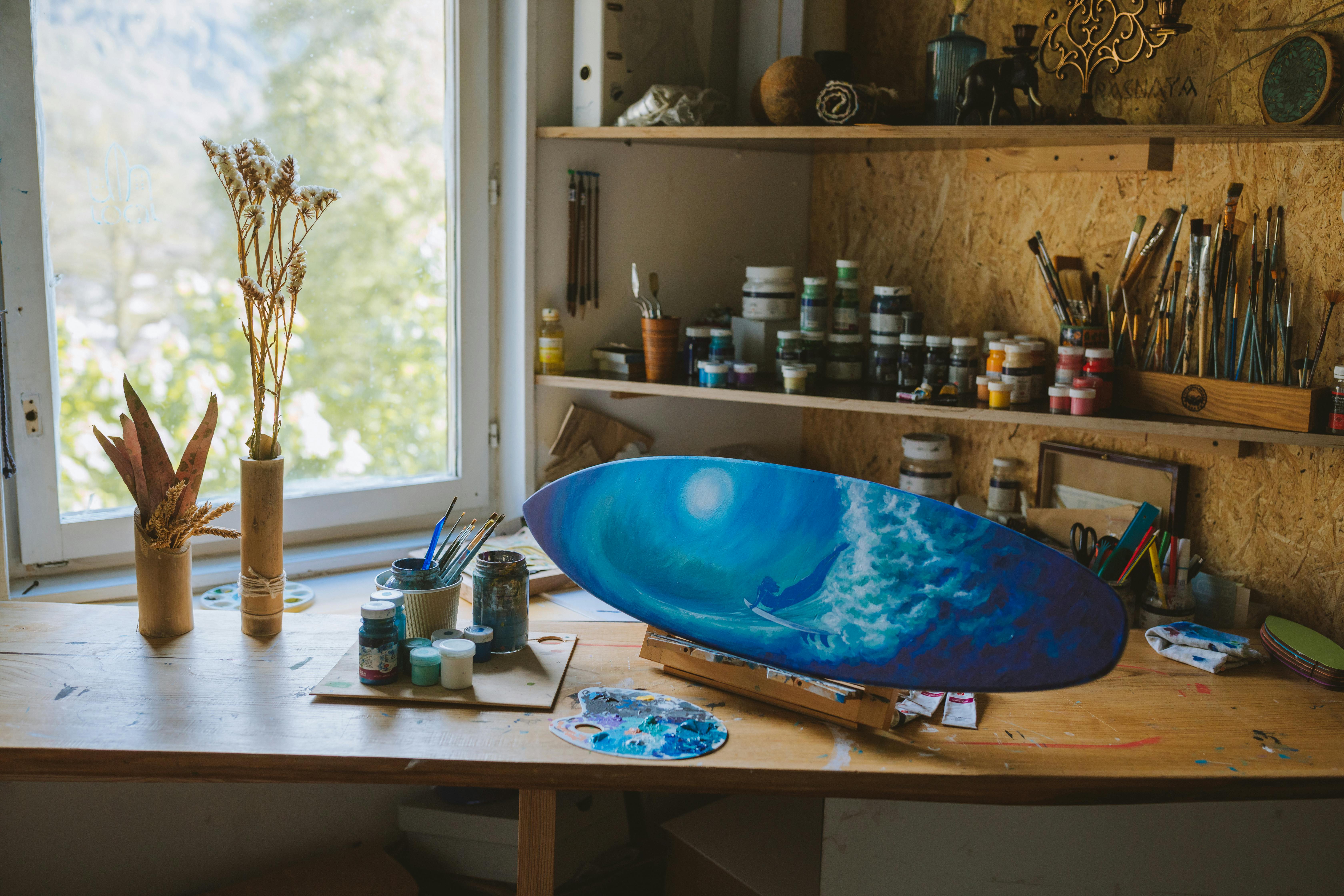 Free An artistic home studio featuring a vibrant blue ocean painting on a skateboard with art supplies and brushes near a window. Stock Photo