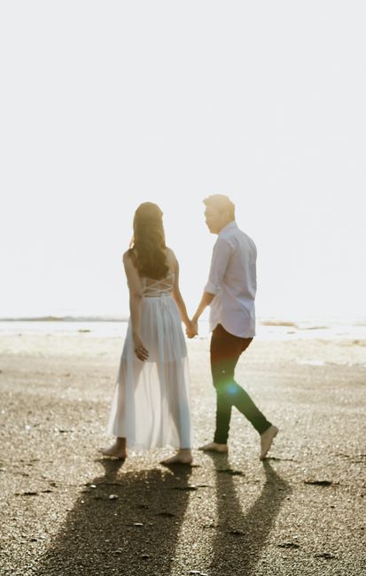A couple holding hands and walking on a sunny beach, captured in a bright, romantic setting.